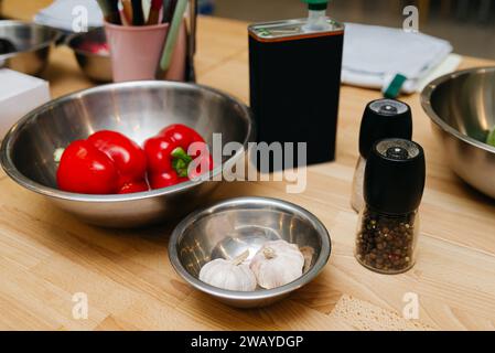 Red bell peppers and whole garlic cloves in stainless steel bowls with a pepper grinder, all set on a wooden countertop for meal preparation. Stock Photo