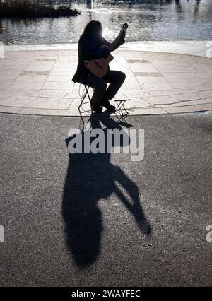 A street musician playing a guitar in the bright Winter sunshine casts a shadow on the ground performing alongside the river Avon in Stratford upon Av. Stock Photo
