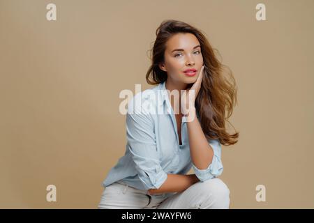 Dreamy woman in light blue shirt leaning on hand, beige background Stock Photo