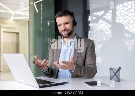 Smiling young man sitting at desk in office wearing headset and talking gesturing hands on video call via laptop. Stock Photo