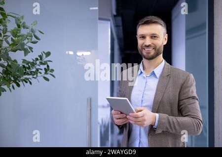 Portrait of a smiling young businessman standing in the lobby of an office center, holding a tablet and looking at the camera. Stock Photo