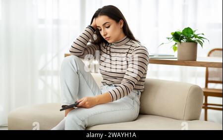 A contemplative young woman in a striped top and jeans lounges on a sofa, phone in hand Stock Photo