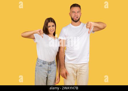 Disapproving young couple in white t-shirts giving thumbs down gestures with displeased expressions Stock Photo