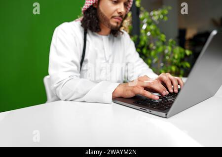 Young Arab man in traditional dress thobe working on computer in office Stock Photo