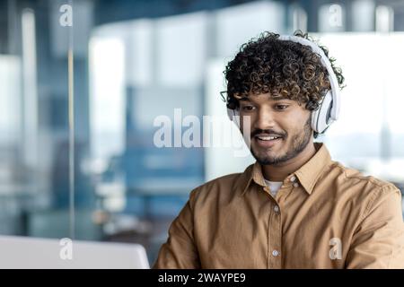 Young indian programmer working inside office with laptop, man in headphones coding new software, businessman developer satisfied with achieved results smiling. Stock Photo