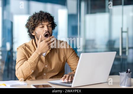 Young overworked and overtired businessman yawning, working inside office with laptop, boring long routine work, man in casual clothes. Stock Photo