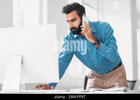 Focused indian man multitasking with phone and computer at desk Stock Photo