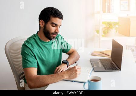 Focused man with beard and green t-shirt writing notes in a notebook Stock Photo