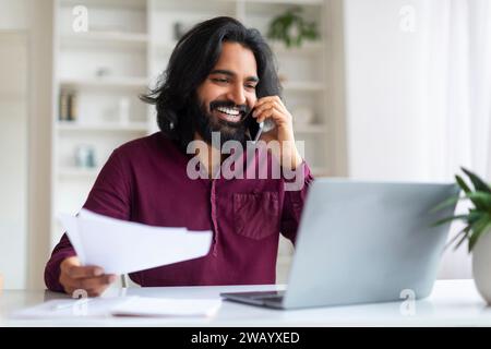 Indian man discussing work on phone while reviewing documents and using laptop Stock Photo