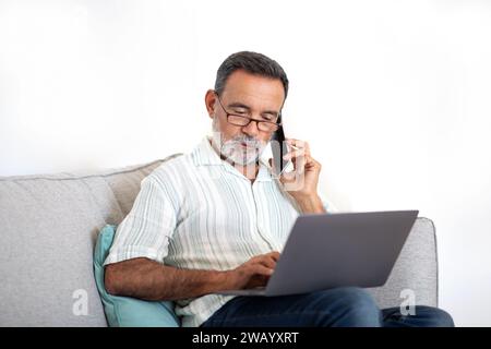 Senior Man Talks on Cell Phone Working On Laptop Indoors Stock Photo