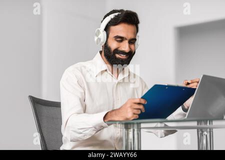 Happy indian man in headphones using tablet and laptop at modern office desk Stock Photo