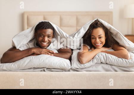 Happy Black Man And Woman Lying Under Blanket In Bed Stock Photo