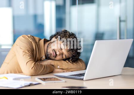 Businessman sleeping at workplace, young overtired and overworked man lying on desk inside office with laptop, long work until late, deadline in evening. Stock Photo