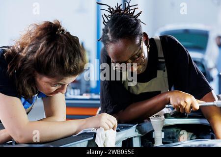 BIPOC mechanics collaborating on servicing broken vehicle, checking for faulty radiator. Women in auto repair shop working together on fixing car, discussing best options Stock Photo