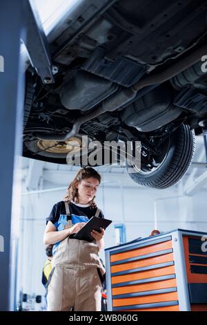Certified mechanic in garage using laptop to follow checklist while doing maintenance on car. Precise employee in auto repair shop does checkup on vehicle helped by device Stock Photo