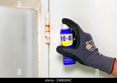 A gloved hand holds a can of WD 40 lubricant lubricating oil spray near a rusty door hinge. Stock Photo