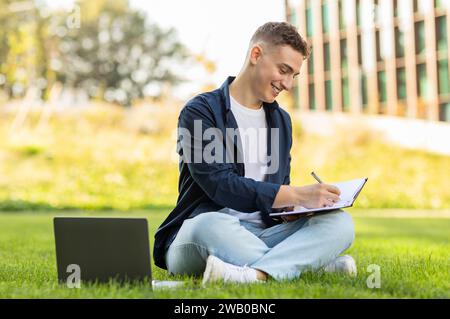 A focused young man sitting on the grass in a park, writing in a notebook with a laptop Stock Photo