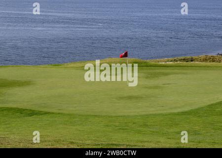 A red and black flag on a white stick in a golf hole along the water. The lush green grass is dipped and raised with its lowest point in the middle. Stock Photo