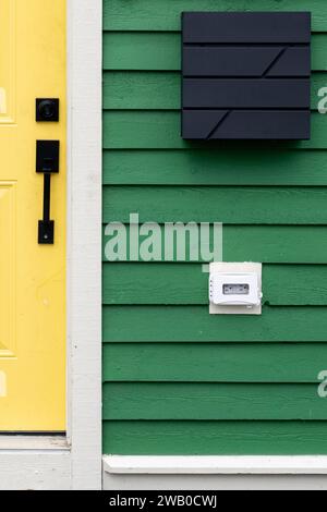 A vibrant yellow colored solid metal door with a black handle and deadbolt lock on a green wooden clapboard style house. There's a modern black square Stock Photo