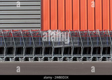 A long row of new metal shopping carts with red handles. The retail pushcarts are in front of an orange and grey colored metal outside wall. Stock Photo