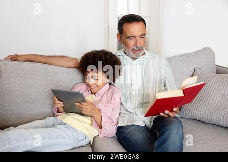 Senior Man Reading Book While Grandson Using Digital Tablet Indoor Stock Photo