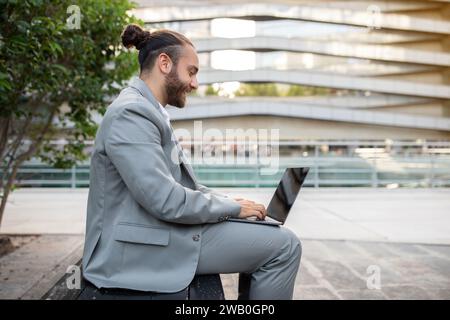 Contented modern businessman with topknot working on laptop outdoors Stock Photo