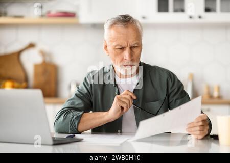 Pensive senior man holding paperwork, focused on reviewing documents in bright kitchen Stock Photo