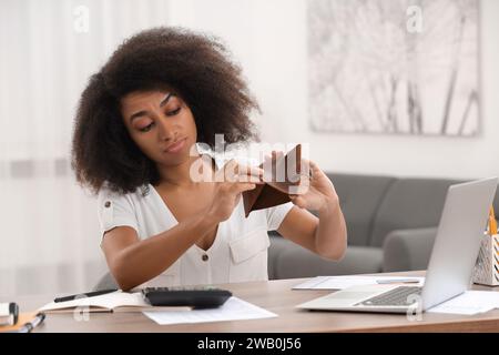 Confused woman with empty wallet planning budget at table in room. Debt problem Stock Photo