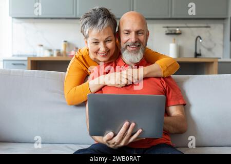 Senior Spouses Using Laptop, Reading Online News On Couch At Home Stock Photo