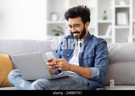 Engrossed indian man in eyeglasses multitasking with laptop and smartphone at home Stock Photo