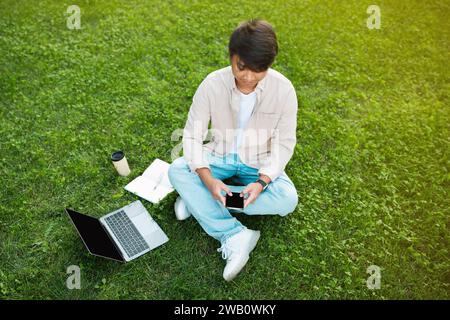 Asian man sitting on grass while using phone and laptop Stock Photo
