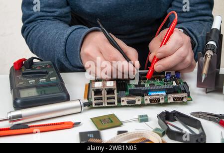 Close-up of a technician's hands in a workshop. The repairer is using a multimeter to measure the parameters of an electronic device on a table. Stock Photo