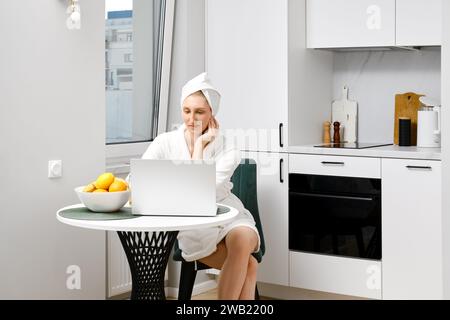 Serious young woman in bathrobe sits in the kitchen with laptop Stock Photo