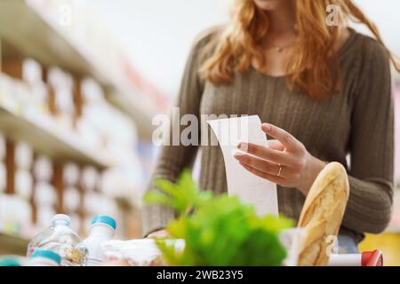 Woman pushing a full shopping cart at the supermarket and checking a long grocery receipt, budget and expense concept Stock Photo