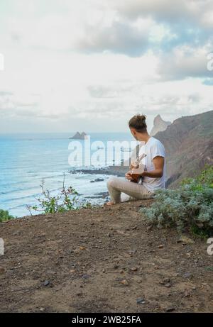 A tranquil scene of face less young man engrossed in playing the guitar, seated on a cliff with the vast ocean and rocky outcrops in the backdrop Stock Photo
