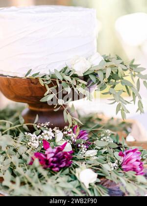 White cake on a wood cake stand decorated with greens and florals Stock Photo