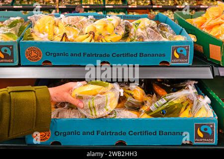 Fair trade plastic bagged organic Bananas cardboard boxes shoppers hand self service fruit in Tesco supermarket shelves in large food store England UK Stock Photo