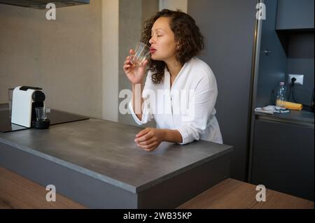 Young adult Hispanic woman in a white robe taking medication, drinking water, standing at the kitchen counter. Pretty female drinks water to renew the Stock Photo