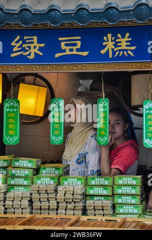 A Chinese Muslim lady and a shy girl selling traditional green bean cakes through a small shopfront in the Muslim Quarter, Xi'an, Shaanxi, China Stock Photo