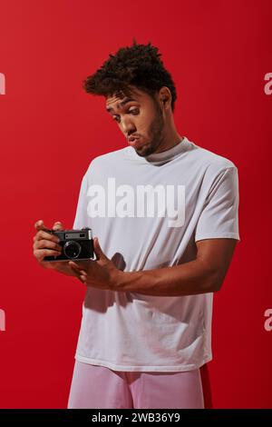 young african american man looking at his vintage camera on red background, photography as hobby Stock Photo
