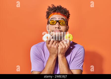 funny young african american man in sunglasses holding bite-sized donuts on orange background Stock Photo