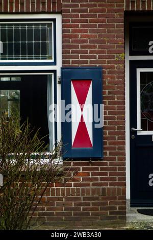 A blue wooden shutter with a red diagonal cross mounted on a brick wall next to a window and a door. Stock Photo