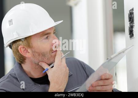 electrician holding clipboard inspecting installation on wall Stock Photo