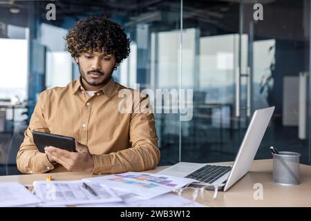 Serious thinking businessman financier with calculator in hands, male accountant reconciles and lists data, paper work worker focused sitting with laptop inside office. Stock Photo