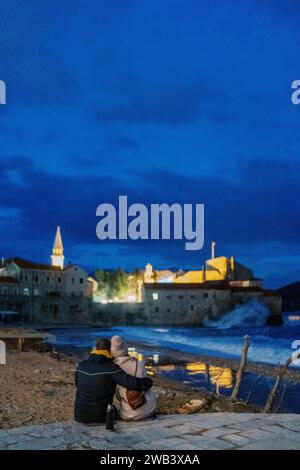 Couple sits embracing on the seashore and looks at the illuminated ancient town. Back view Stock Photo