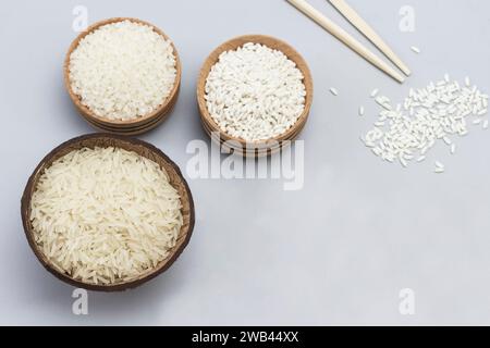 Three types of white rice in wooden bowls and coconut shells. Wooden spoon with rice and sticks. Gray background. Flat lay Stock Photo