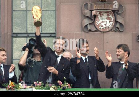 FILED - 09 July 1990, Hesse, FrankfurtMain: The then team manager Franz Beckenbauer (l) holds up the World Cup trophy he won in Italy on the balcony of the Römer, laughing. He is flanked by (from left): Raimond Aumann and Günther Hermann. Many thousands of people give the players of the German soccer World Cup team an enthusiastic welcome on the Römerberg. The German national soccer team is world champion for the third time. The previous evening, Argentina had been defeated 1:0 in the final in Rome. Franz Beckenbauer is dead. The German soccer legend died on Sunday at the age of 78, his famil Stock Photo