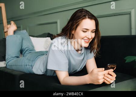 Sociable Relaxation. Woman Typing, Socializing on Smartphone While Lounging. Digital Socialization. Happy Woman Typing, Using Smartphone on Couch Stock Photo