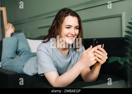 A cheerful young woman typing on her smartphone, using social media, socializing, lying on the couch. Connected and Content. Young Woman Engages on Stock Photo