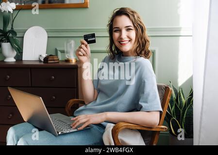 Young Lady Excitedly Using Credit Card for Online Purchase. E-commerce Enthusiasm. Smiling Woman Shows Credit Card with Laptop. Digital Shopping Stock Photo